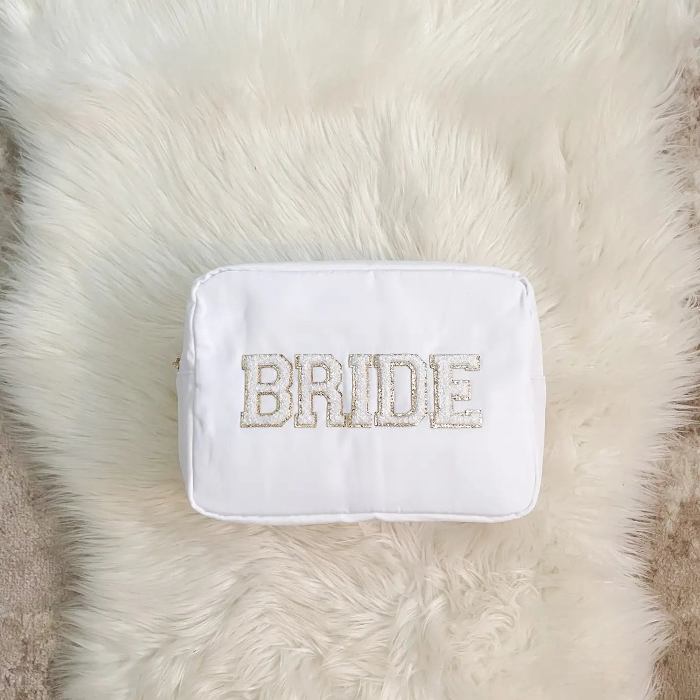 White nylon makeup bag with “BRIDE” patch lettering in white and gold in size extra large, placed on a soft ivory faux fur rug.