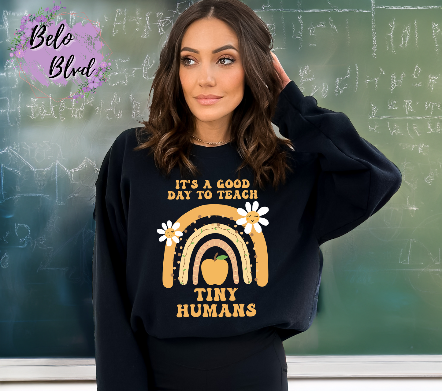 Woman in black crewneck sweatshirt with cheerful teacher-themed graphic in gold and white, standing in front of a chalkboard.
