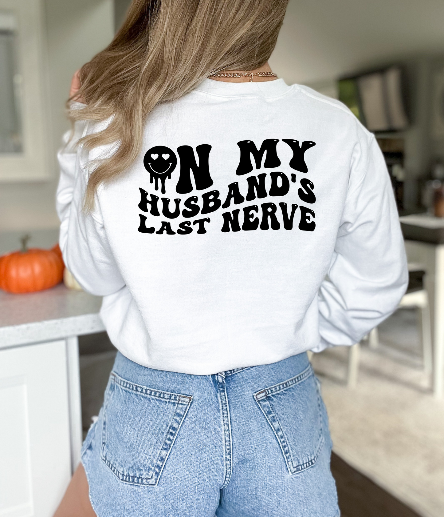 Back view of a woman wearing a white crewneck sweatshirt with the bold text "On My Husband’s Last Nerve" and a smiley face with heart eyes printed on the back.