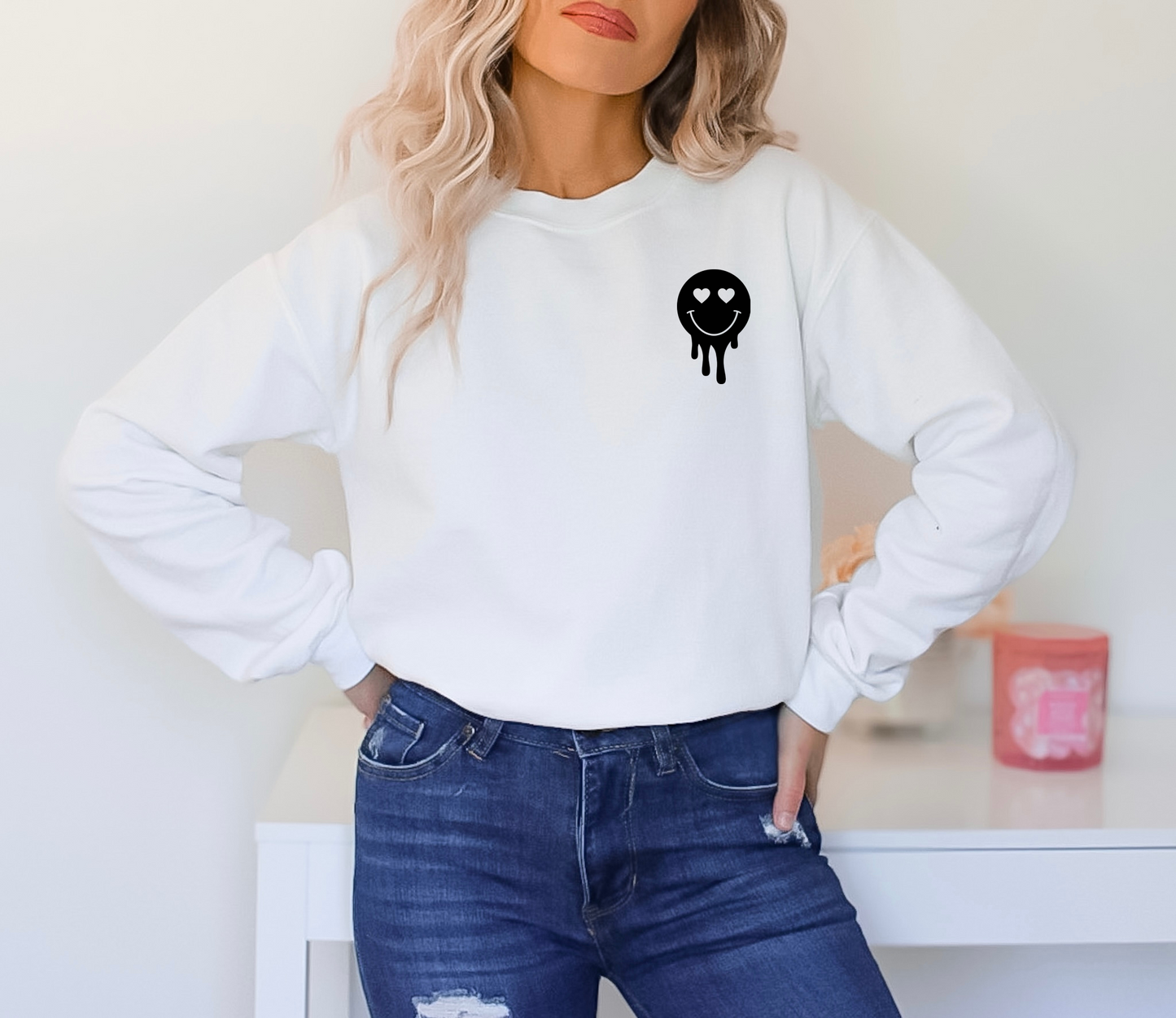 Front view of a woman wearing the white crewneck sweatshirt showing a small black dripping smiley face with heart eyes on the left chest.