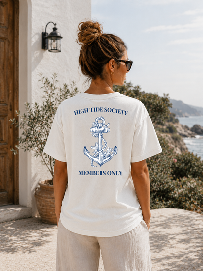 Person wearing a white t-shirt with an anchor design and text, standing outdoors near a building and ocean.