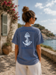 Person wearing a blue t-shirt with an anchor design and text, standing by a scenic waterfront.