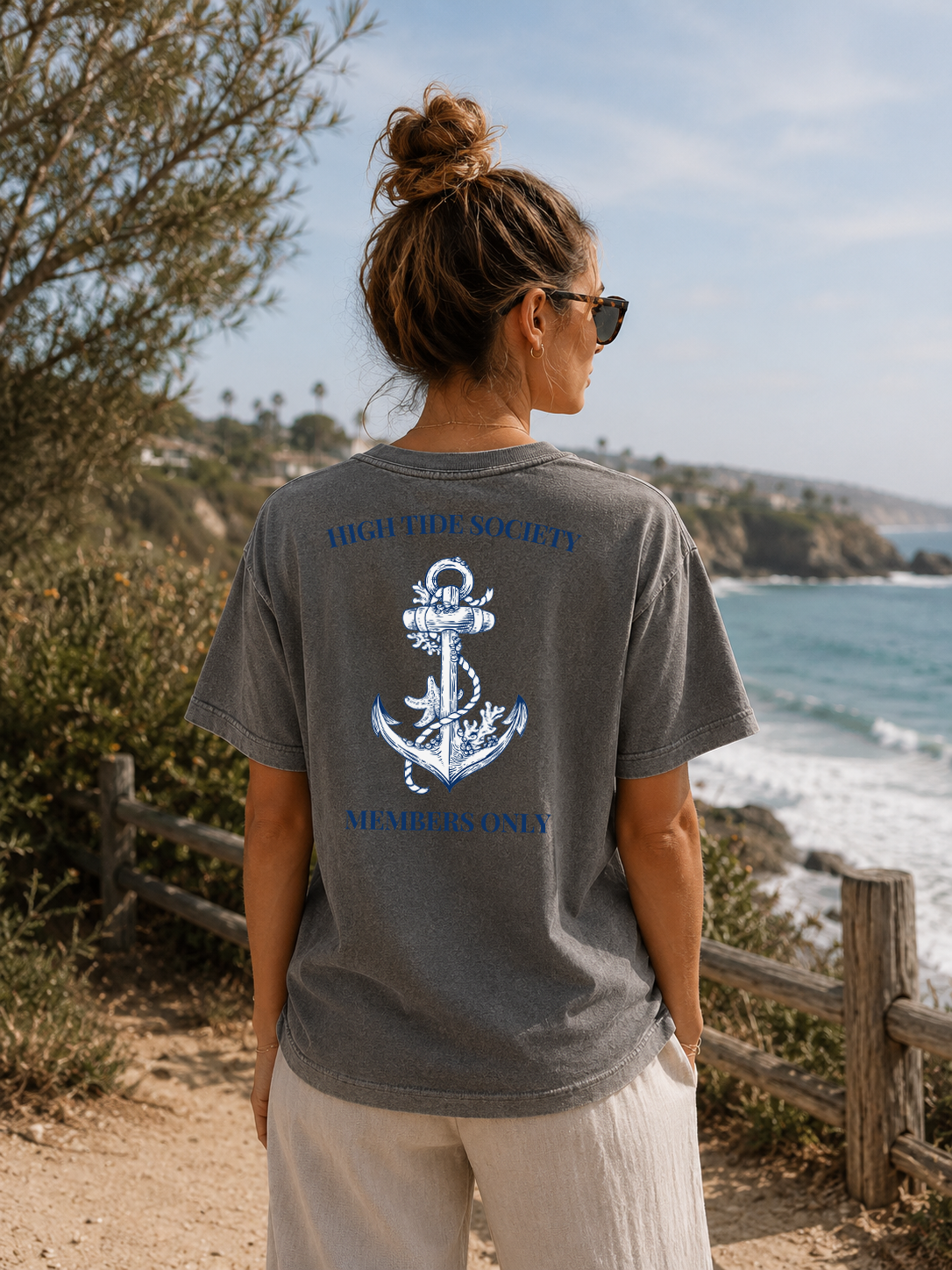 Person wearing a gray t-shirt with an anchor design and text, standing by a scenic coastal area.