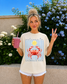 Woman wearing a 'La Dolce Vita' t-shirt with a red crab design, holding a smoothie and making a peace sign.