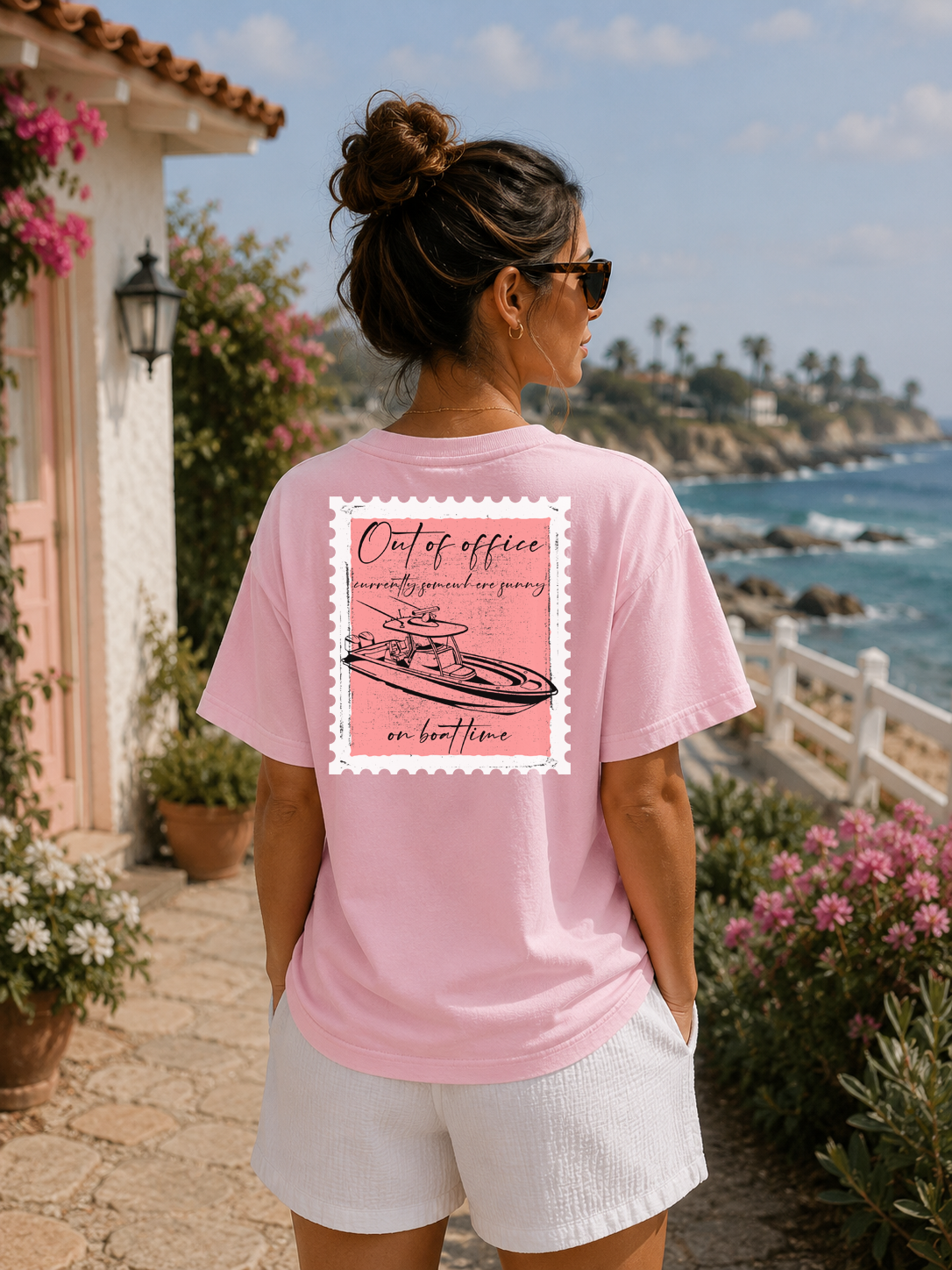 Woman wearing a pink t-shirt with a graphic design, standing by a scenic coastal area.