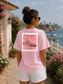 Woman wearing a pink t-shirt with a graphic design, standing by a scenic coastal area.