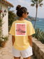 Woman wearing a yellow t-shirt with a design on the back, standing by a scenic coastal area.