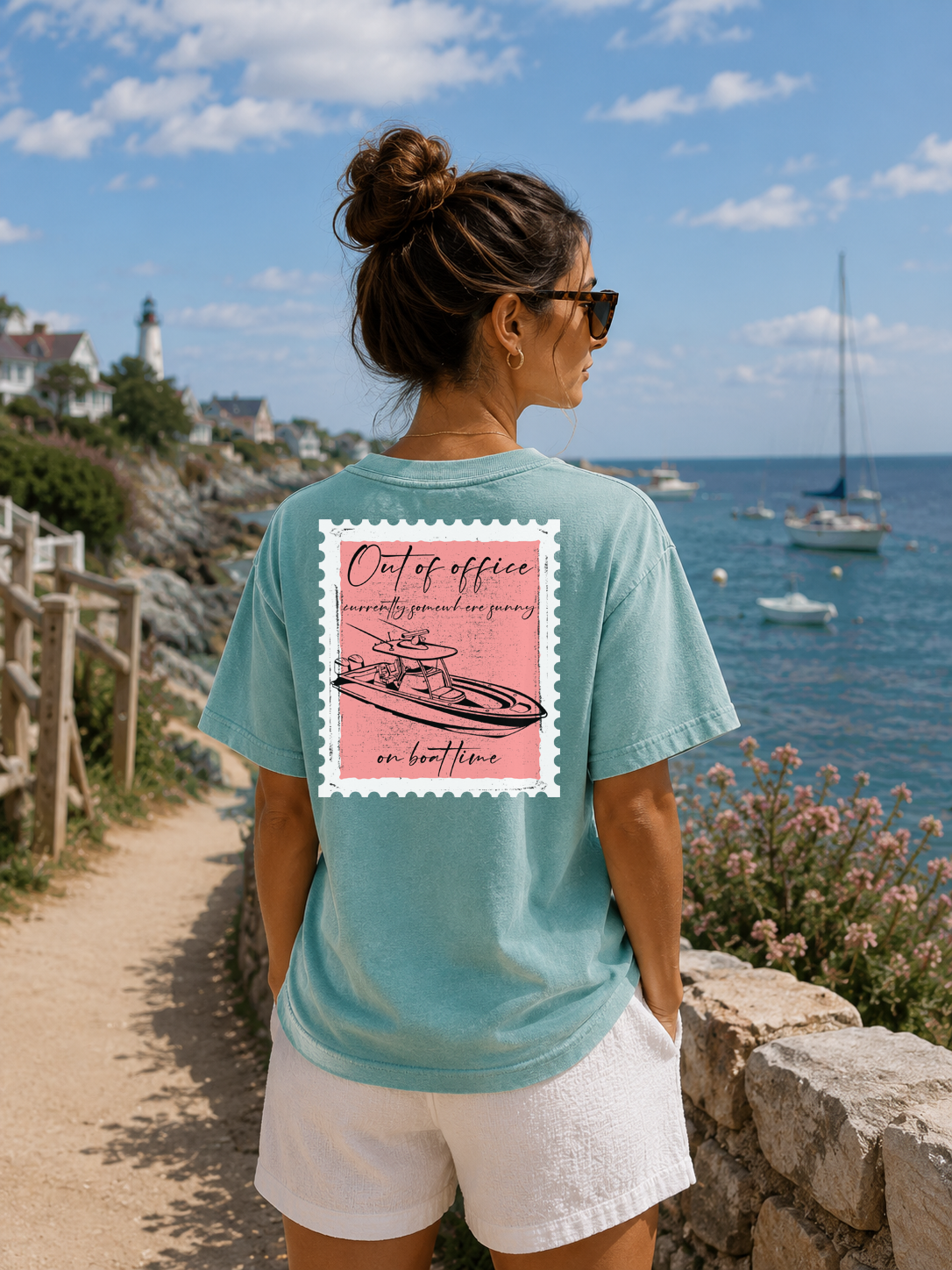Woman wearing a teal t-shirt with a pink graphic and text, standing by a scenic coastal view.