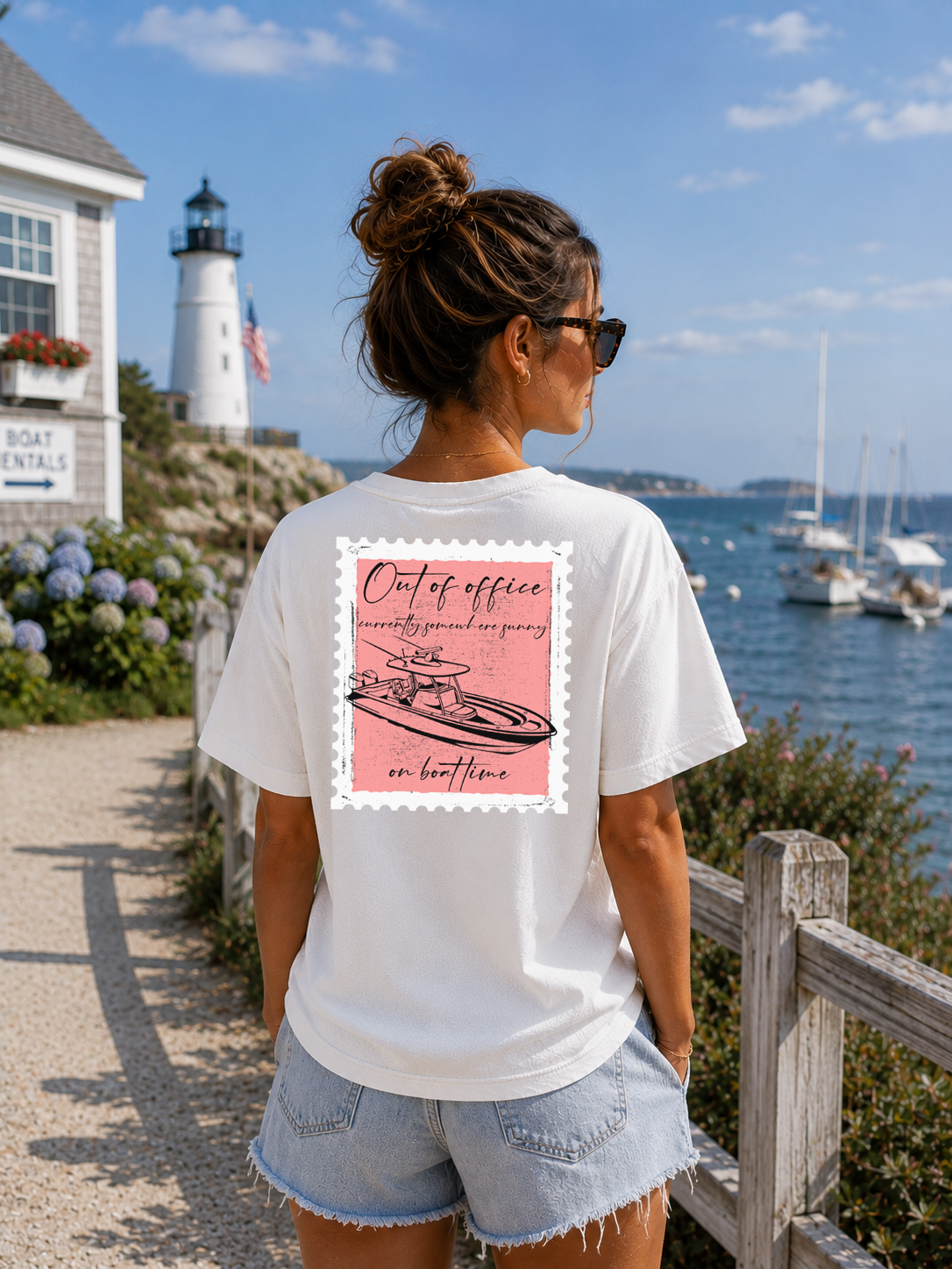 Woman wearing a white t-shirt with a graphic design, standing by a lighthouse and waterfront.