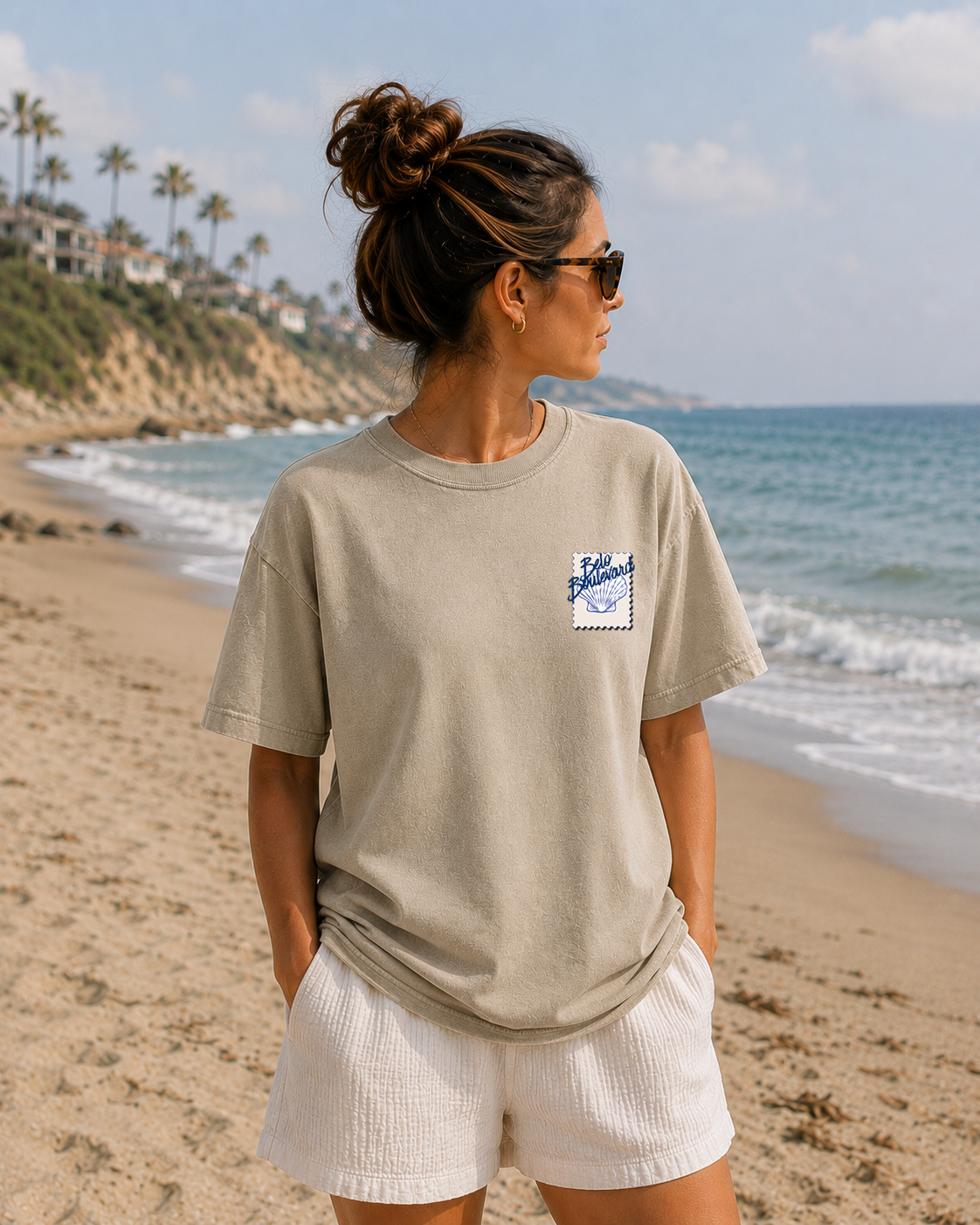Woman standing on a beach wearing a beige t-shirt with a logo and white shorts, looking out towards the ocean.