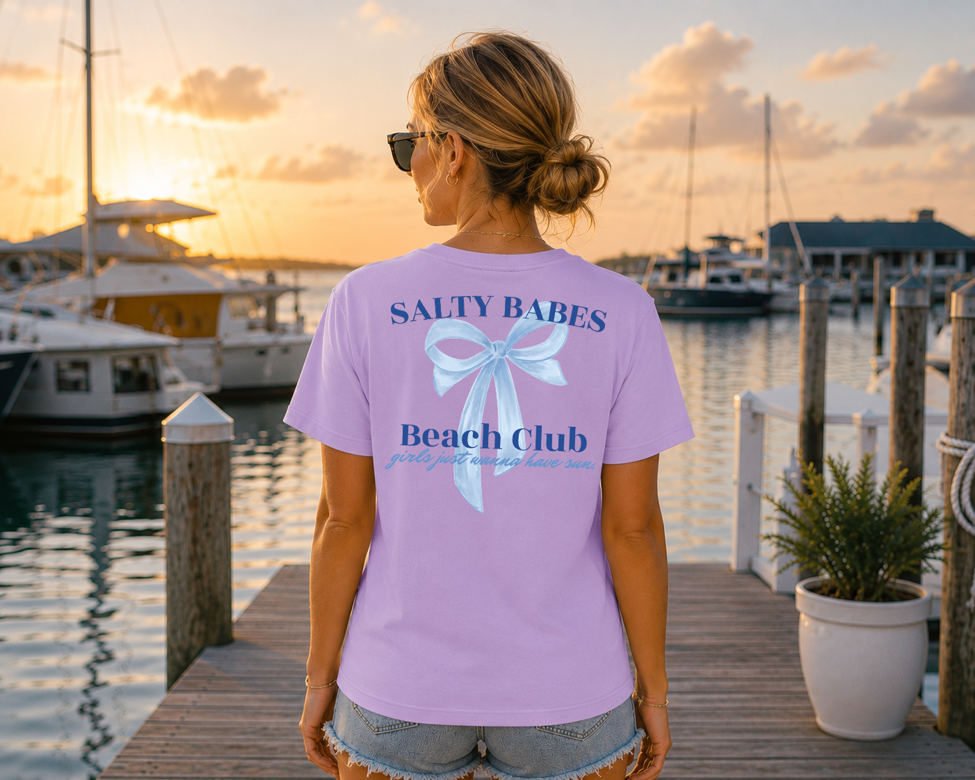 Woman wearing a 'Salty Babes Beach Club' shirt by a marina at sunset.