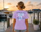 Woman wearing a 'Salty Babes Beach Club' shirt by a marina at sunset.