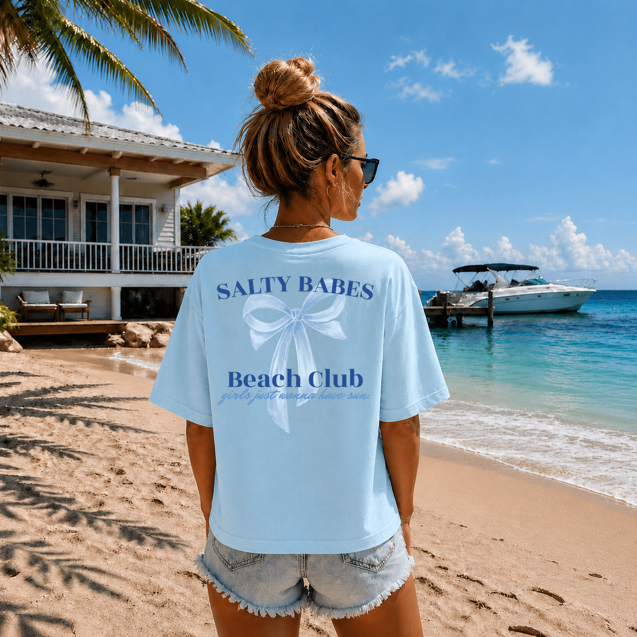 Woman wearing a 'Salty Babes Beach Club' t-shirt on a beach with a house and boat in the background.