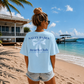 Woman wearing a 'Salty Babes Beach Club' t-shirt on a beach with a house and boat in the background.