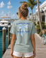 Woman wearing a 'Salty Babes Beach Club' t-shirt by a marina with palm trees and a building in the background.
