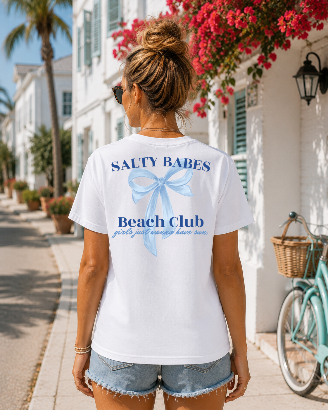 Woman wearing a 'Salty Babes Beach Club' t-shirt on a sunny street with flowers and a bicycle.