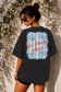 Woman wearing a black t-shirt with a 'Sea Breeze Please' design against a floral wall.