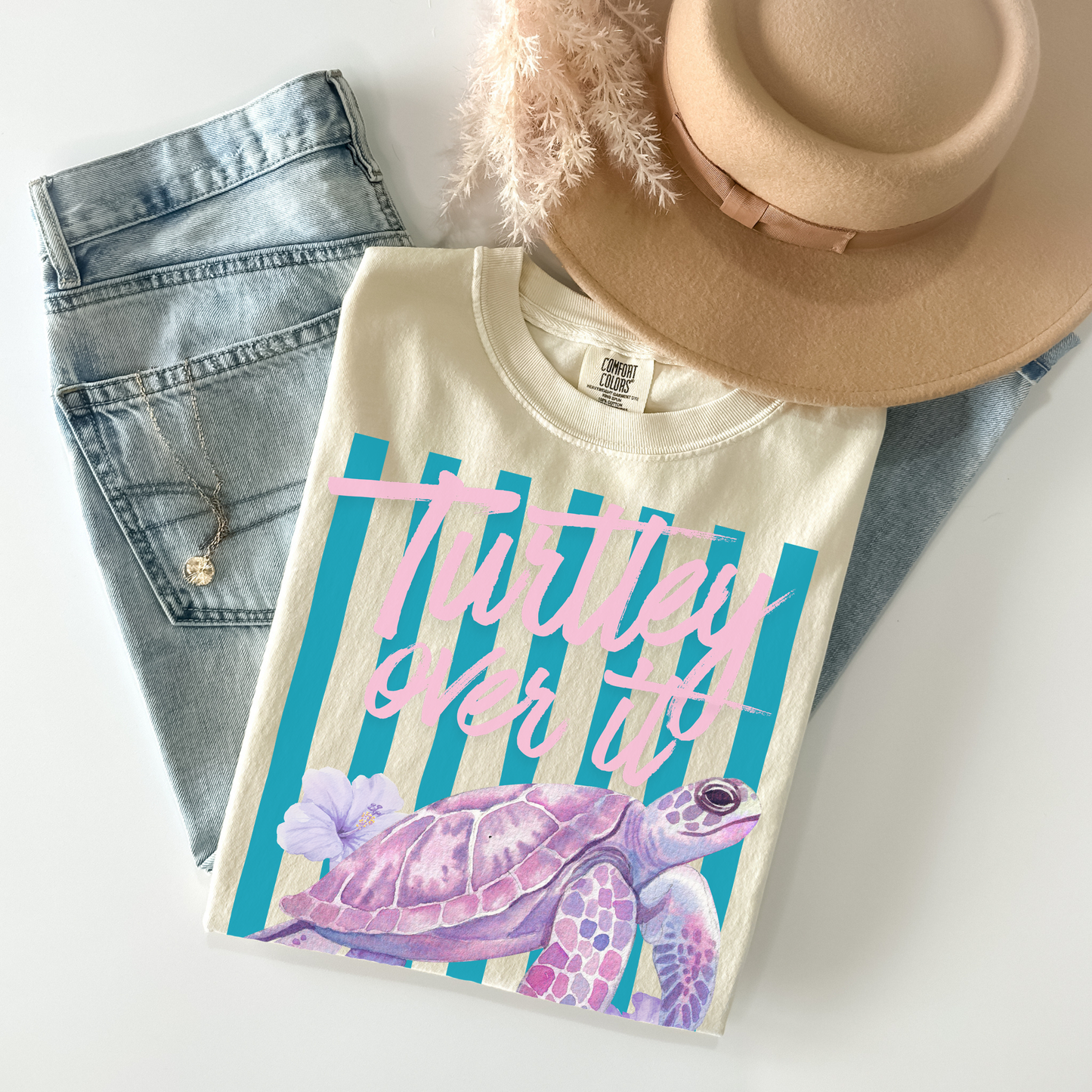 T-shirt with a turtle design and text, placed on denim jeans and a beige hat.