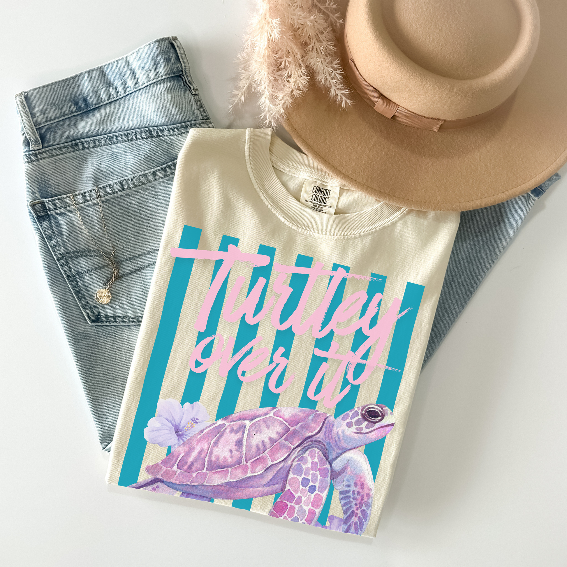 T-shirt with a turtle design and text, placed on denim jeans and a beige hat.