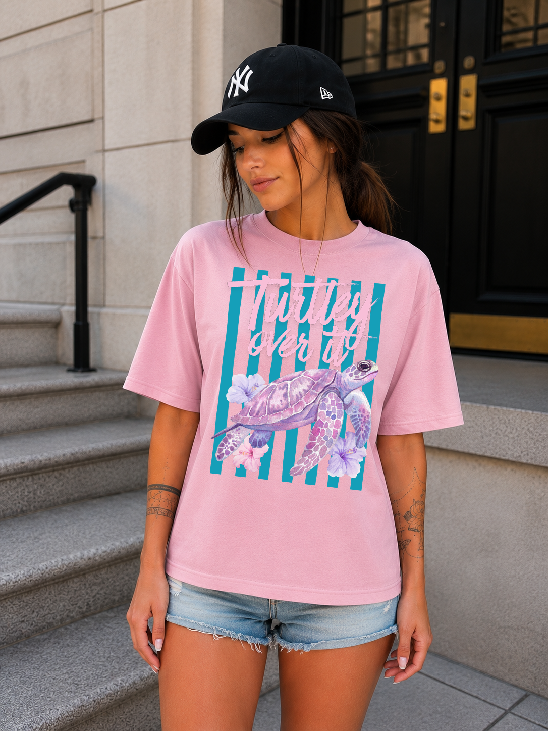 Woman wearing a pink t-shirt with a turtle graphic and a black cap, standing on steps.