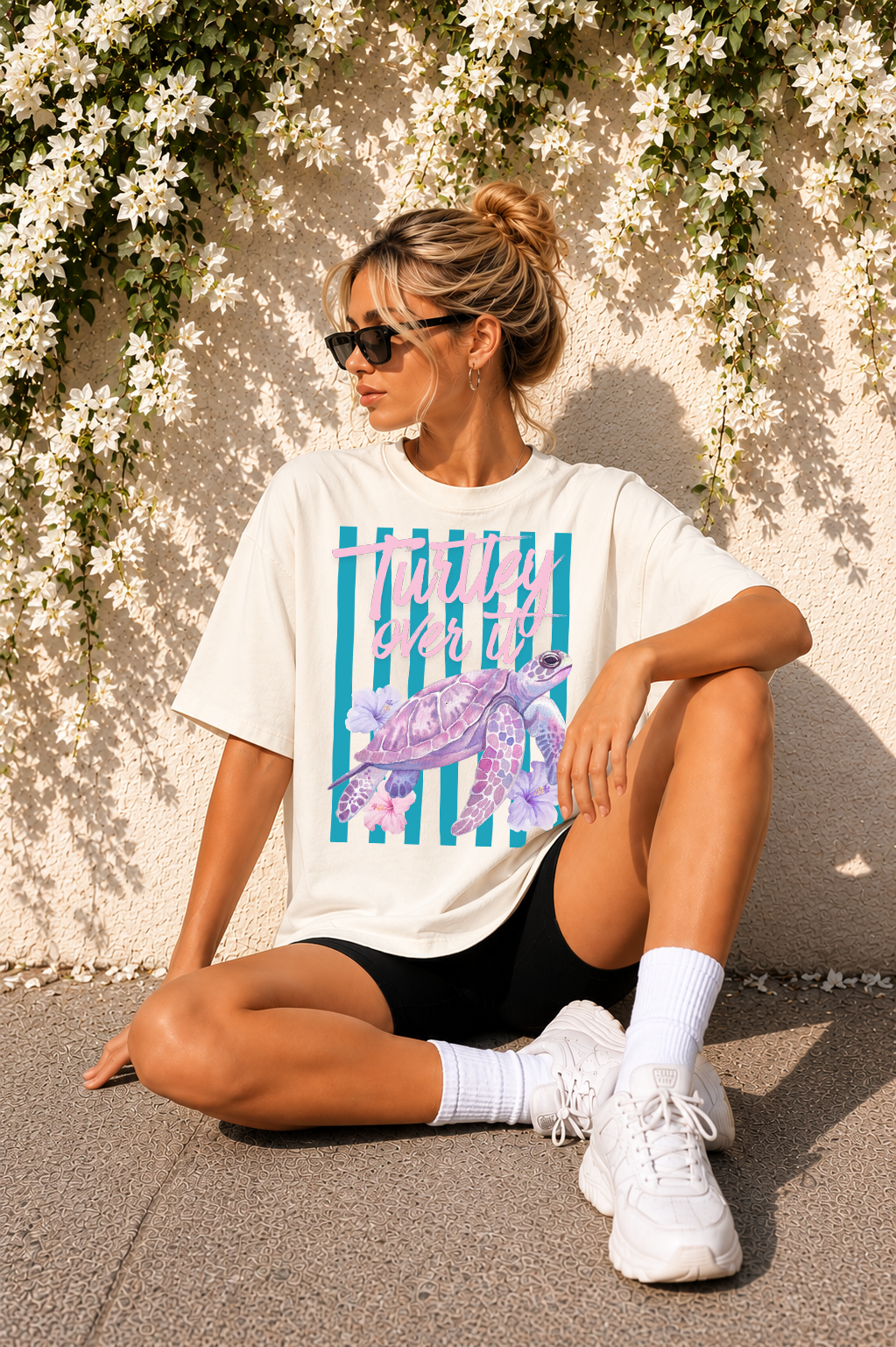 Woman sitting on the ground wearing a graphic t-shirt with a turtle design, black shorts, white socks, and white sneakers.