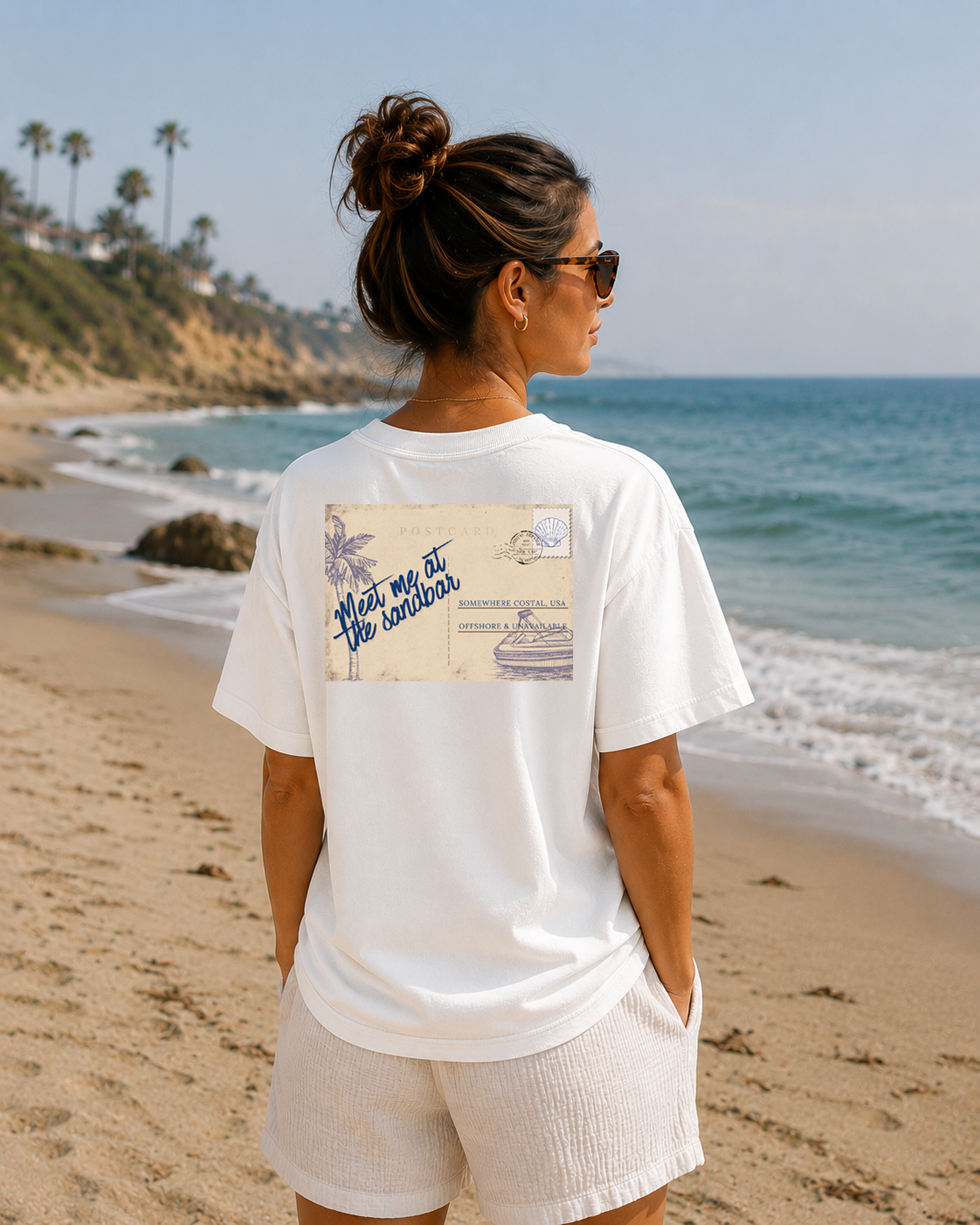 Woman standing on a beach wearing a white t-shirt with a graphic design, facing the ocean.