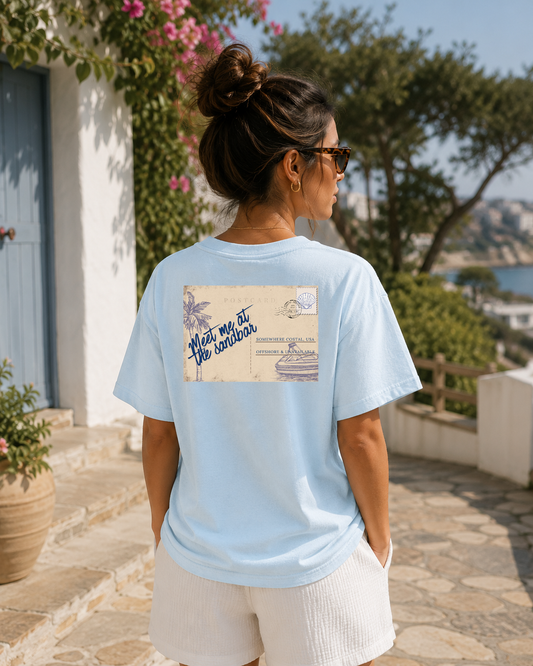 Woman wearing a light blue t-shirt with a graphic design, standing outdoors near a building with a blue door.