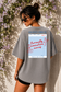 Woman wearing a gray t-shirt with a graphic design on the back, standing against a floral wall.