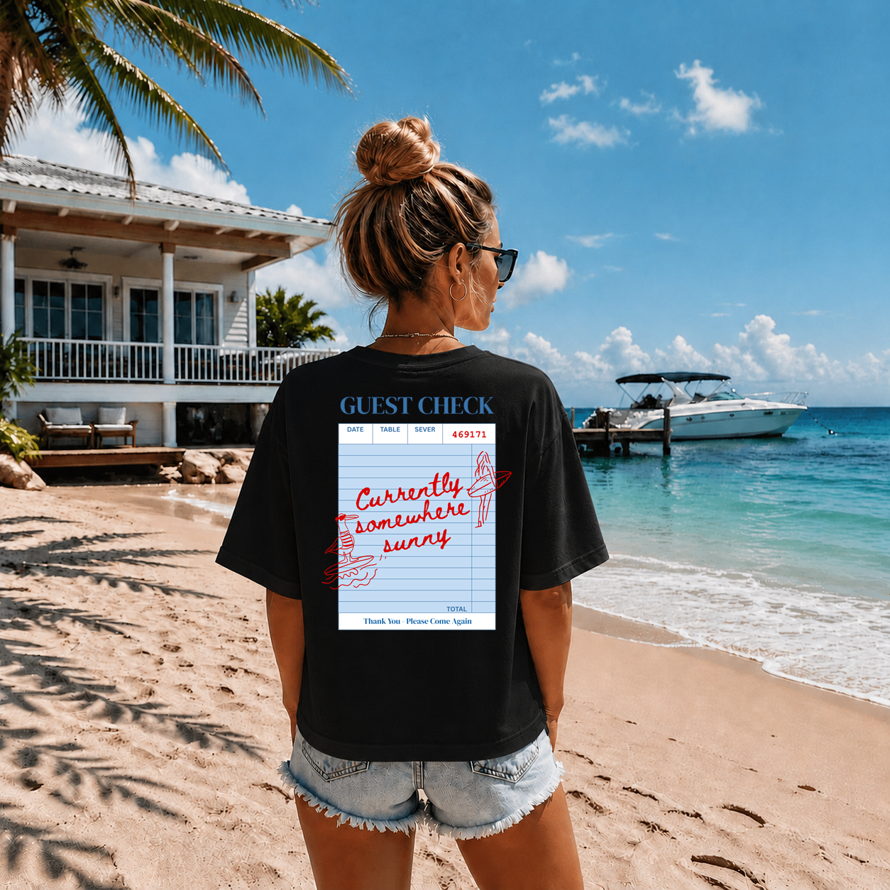 Person on a beach wearing a black t-shirt with a graphic design, looking at a house and boat.